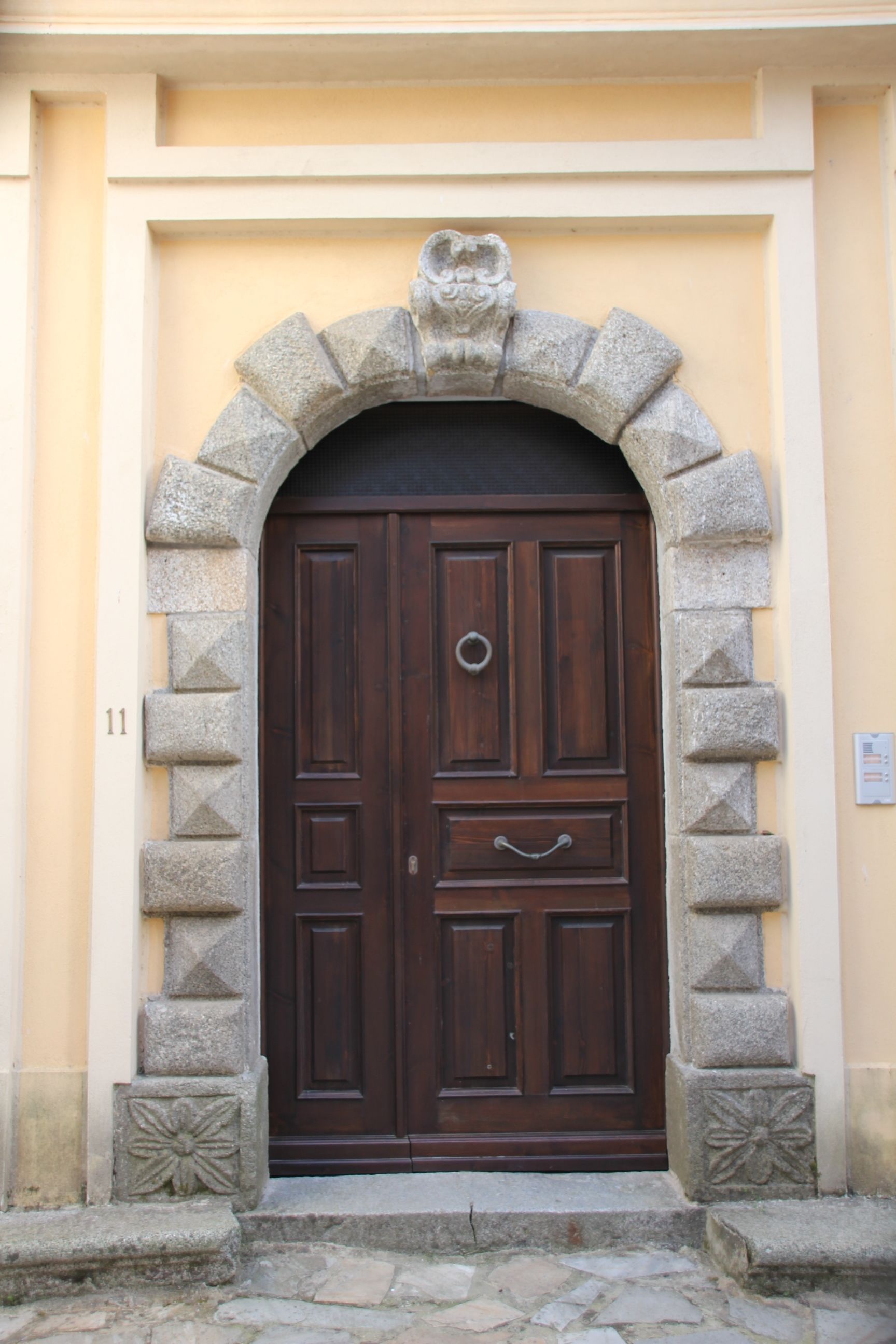 A dark wooden door with a circular handle, flanked by stone pillars with geometric patterns, set within a beige wall with a stone arch above.