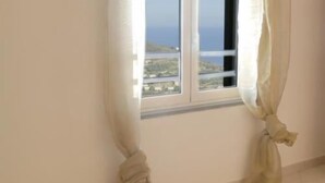 A room with a window and curtains tied back, revealing a view of the sea and buildings.