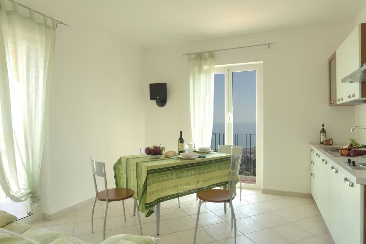 Un coin repas avec une table dressée pour deux personnes, une cuisine équipée d’un évier et de placards, et un balcon avec vue.