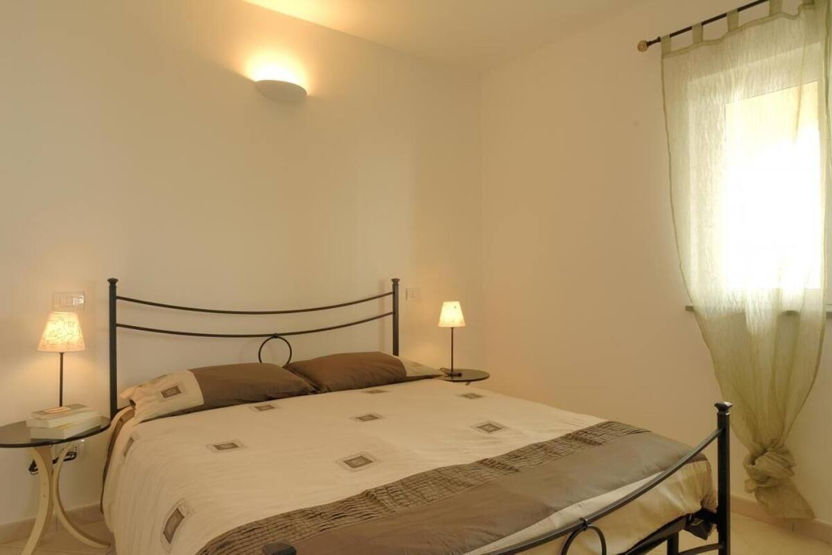 Une chambre à coucher avec un lit, deux lampes de chevet, un luminaire fixé au mur et une fenêtre ornée de rideaux légers.