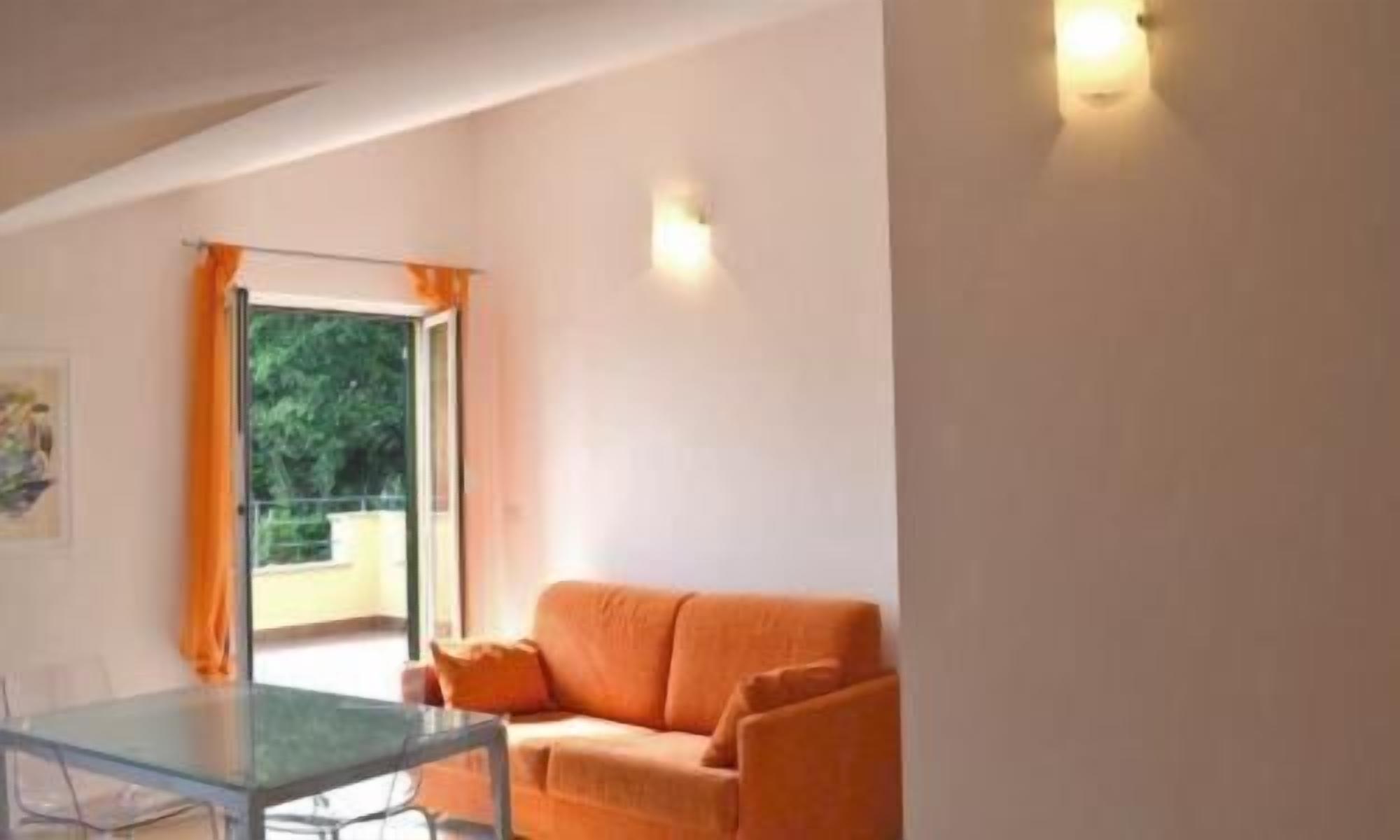 A room with a glass table, orange sofa, and a window with orange curtains.