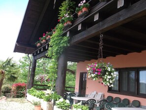 Un área de estar al aire libre techada, con una mesa y sillas, rodeada de plantas en macetas y cestas de flores colgantes.