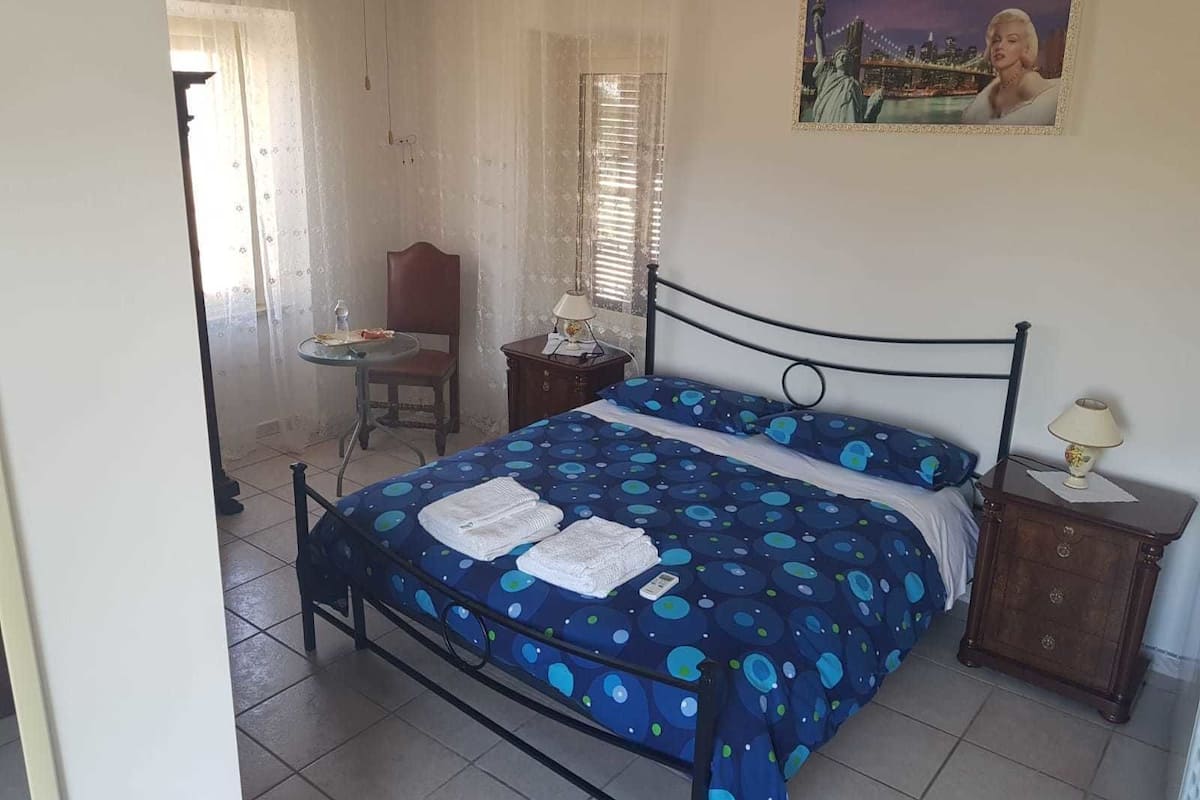 A bedroom with a bed, a chair, a small table, and a nightstand.