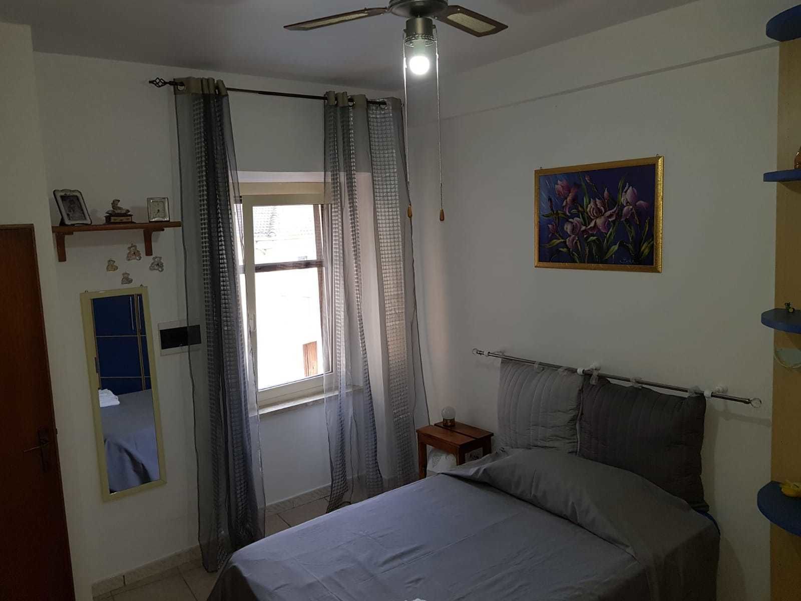 Un dormitorio con cama, cortinas en la ventana, una mesita de noche, un estante en la pared y un cuadro.