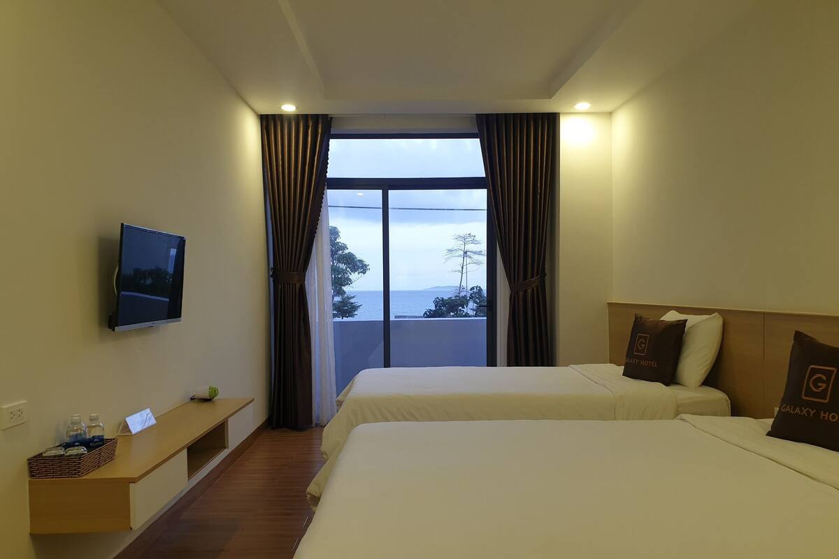 A hotel room with two beds, a TV, a window with curtains, a bedside table with a basket, and a view of the sea.