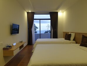 Habitación de hotel con dos camas, televisor, ventana con cortinas, mesita de noche con cesta y vista al mar.