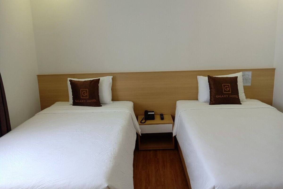 A hotel room with two beds, each with a Galaxy Hotel pillow, a wooden headboard, and a nightstand with a phone and remote control.