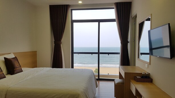 A hotel room with a bed, a TV, a desk, and a view of the ocean. - Galaxy Hotel (Quy Nhon)