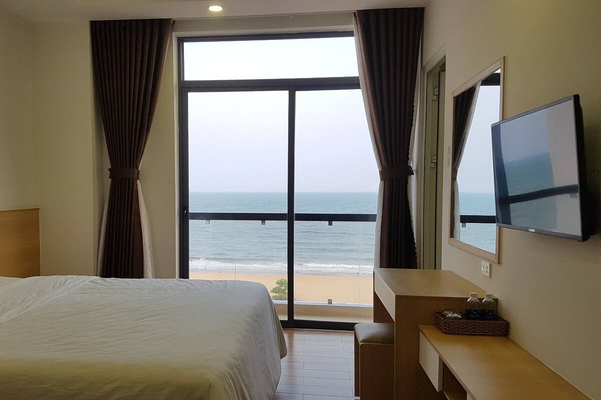 A hotel room with a bed, a TV, a desk, and a view of the ocean.