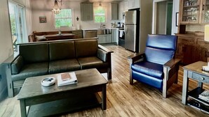 Ein geräumiges Wohnzimmer mit einer Couch, einem Couchtisch und einem blauen Sessel. Im Hintergrund ist eine Küche mit Kühlschrank und Schränken zu sehen.