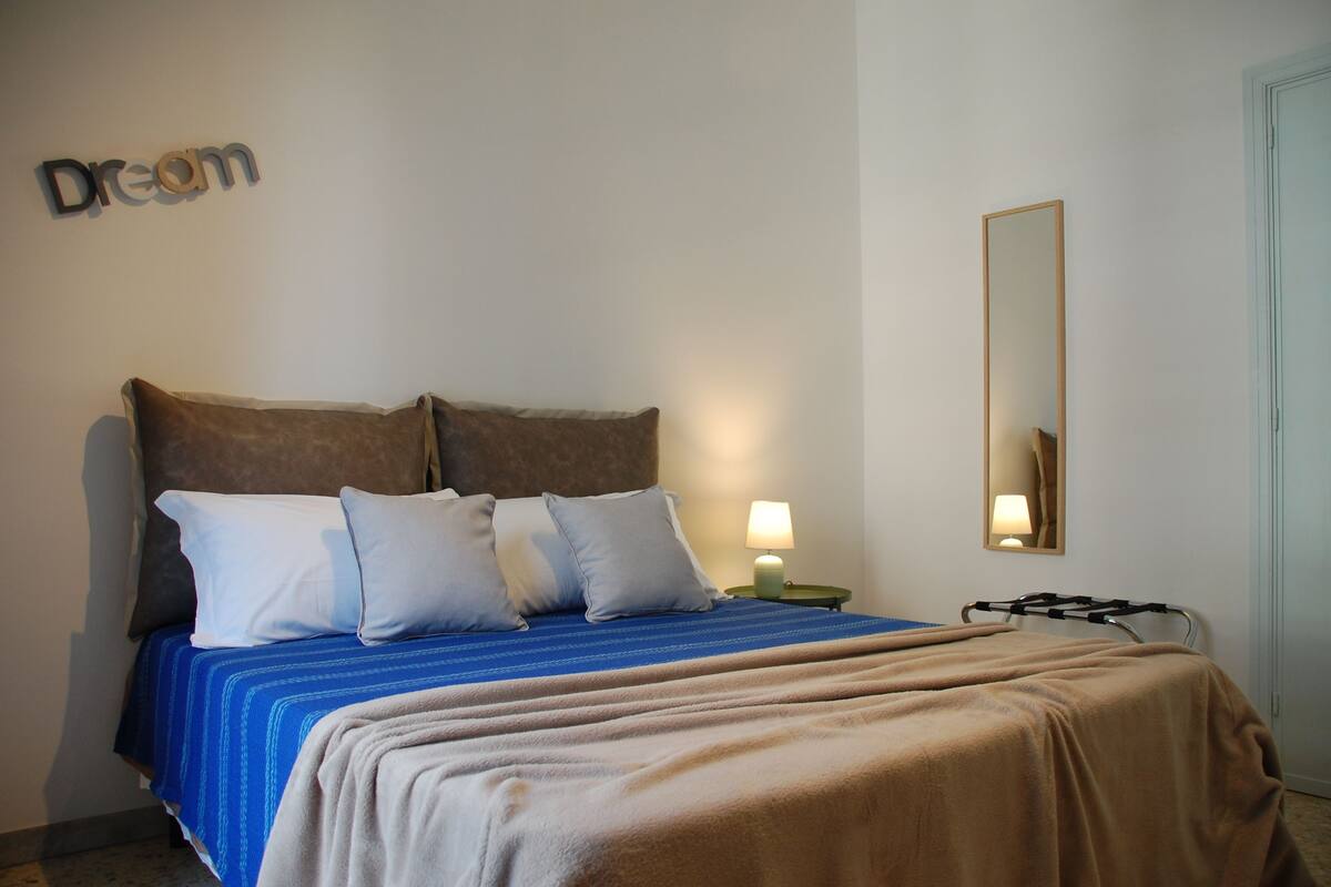 A neatly made bed with a blue and brown bedspread, a small bedside table with a lamp, and a wall-mounted mirror.