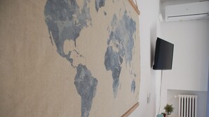 Una habitación con un gran mapa mundial en la pared, un televisor montado, un radiador y una pequeña mesa con una planta y un libro.