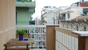 Un balcón con una mesa de madera y una planta en maceta, con vistas a una calle con coches y edificios.