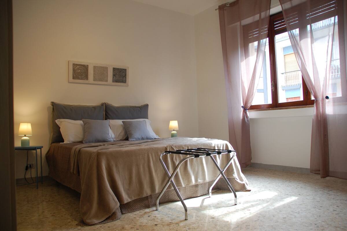 A bedroom with a bed, bedside lamps, a window with curtains, and a wall-mounted artwork.