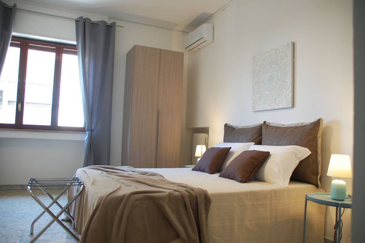 A neatly made bed with a brown blanket, a wardrobe, a wall-mounted air conditioner, and a bedside lamp.