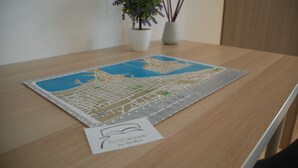 Un mapa detallado de la ciudad sobre una mesa de madera, con una tarjeta pequeña que dice 'BAKINGROOMS' y una planta en maceta con flores moradas.