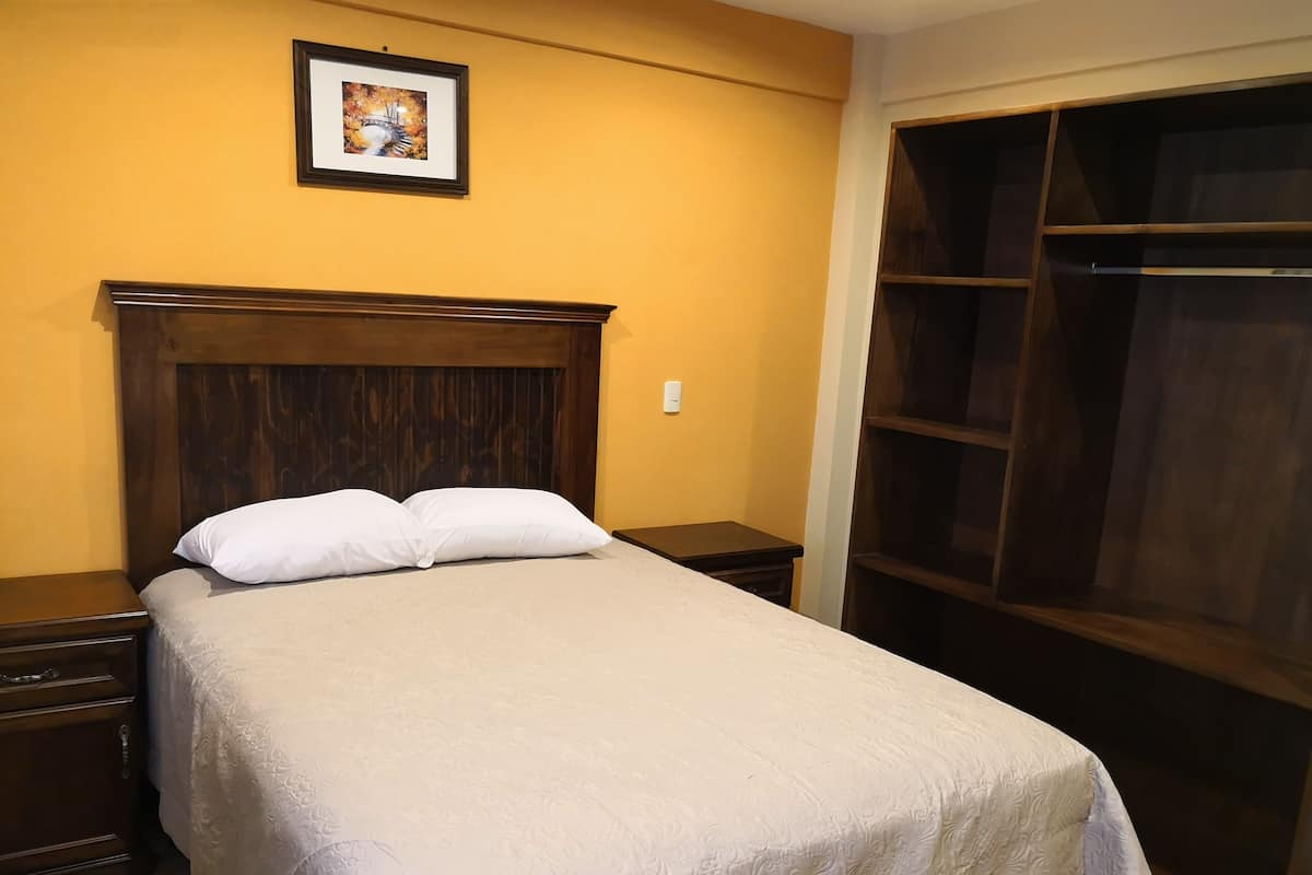 A bedroom with a wooden headboard, a bed with white bedding, a nightstand, a bedside table, a wardrobe, and a framed picture on the wall.