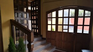 Una escalera de madera con barandilla, macetas con plantas, piso de baldosas y una puerta grande de madera con ventanas.