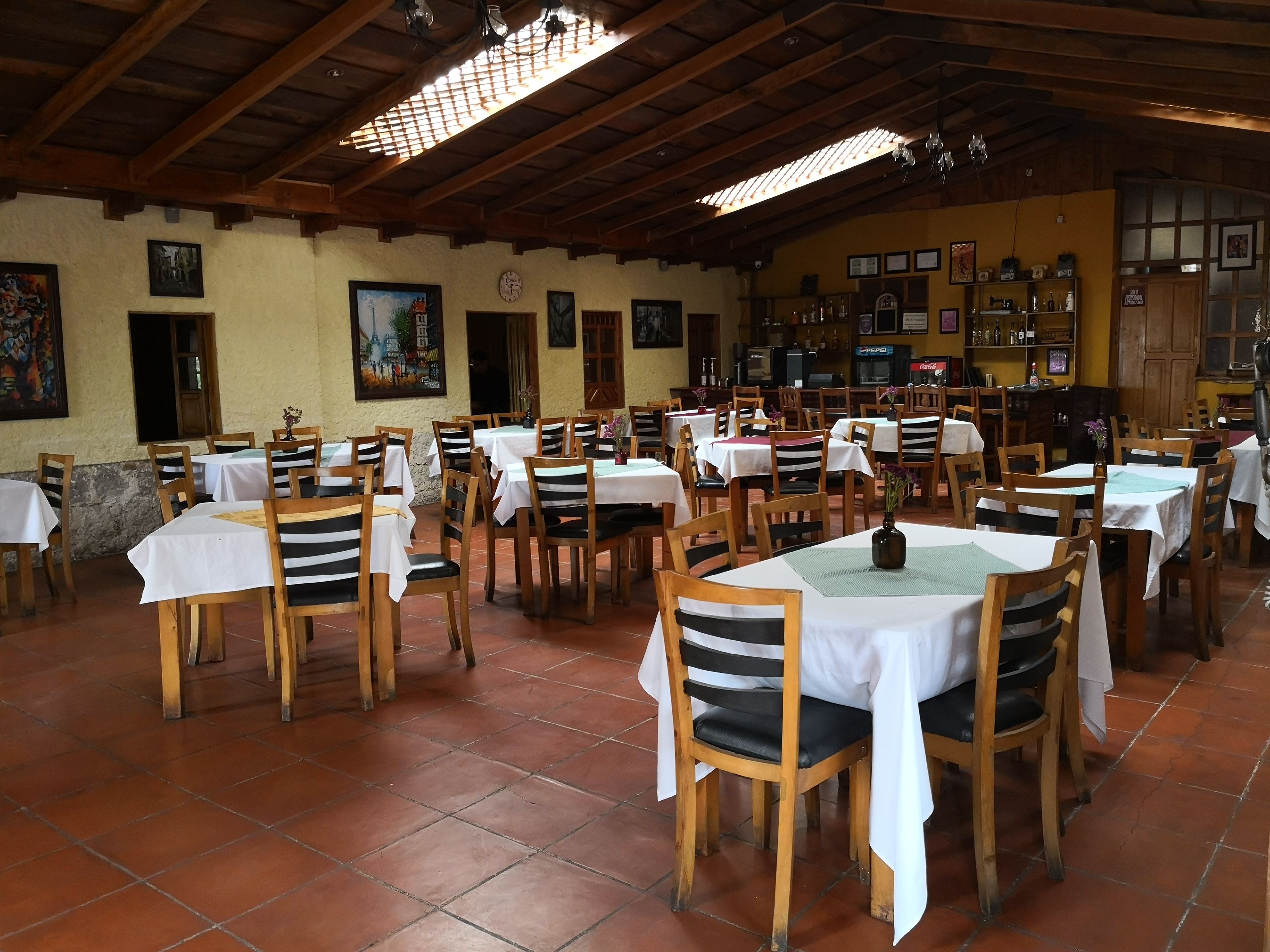 Un restaurante con mesas y sillas de madera, manteles blancos y suelo de baldosas.