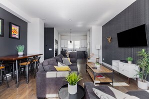 Una sala de estar moderna con un sofá, una mesa de centro y un comedor. Se ve una cocina al fondo con armarios blancos y una encimera negra.