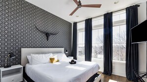 A modern bedroom with a large bed, a ceiling fan, and a view of the outside through large windows.