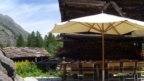 A rustic outdoor seating area with stone tables and chairs, a large umbrella, and a wooden building with a stone roof. - Apartment in Täsch / Zermatt for two persons (Täsch)