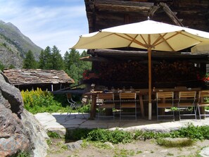 Ein rustikaler Außenbereich mit Steintischen und -stühlen, einem großen Sonnenschirm und einem Holgebäude mit Steinziegeldach.