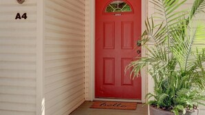 Eine rote Tür mit Fenster, eine Fußmatte mit der Aufschrift 'hello' und ein Blumentopf im Vorbereich.