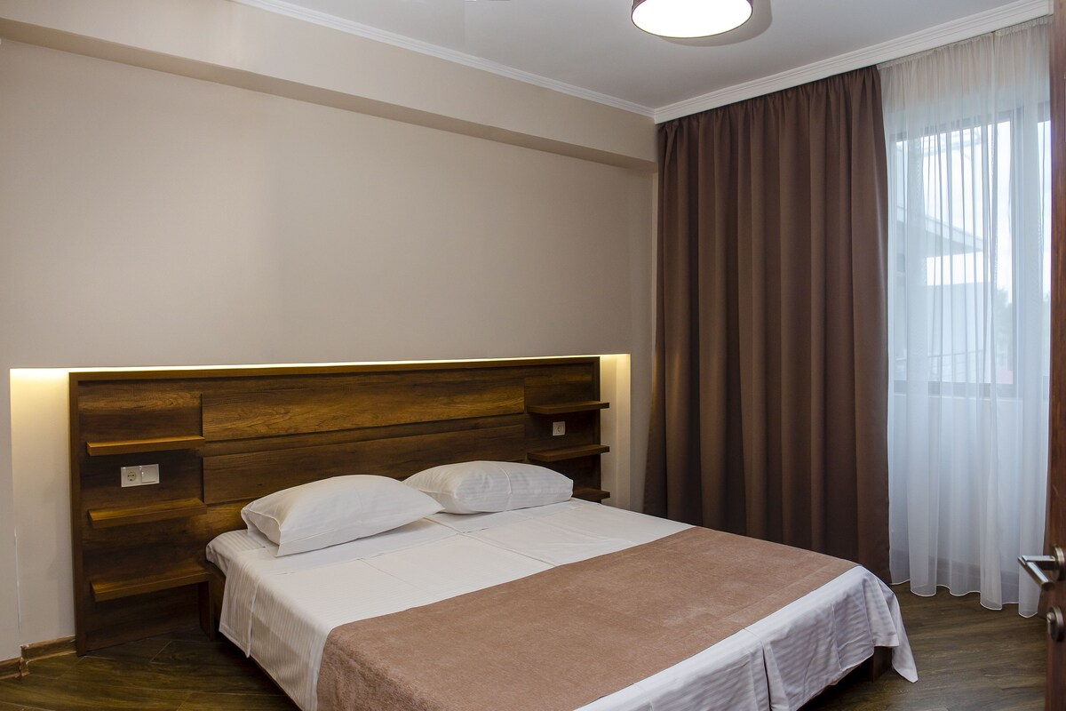 A hotel room with a wooden headboard, a large bed with white linens, a brown throw blanket, and a window with brown curtains.