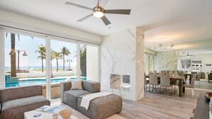 A modern living room with a large window overlooking a pool and beach, a ceiling fan, and a dining area with a marble backsplash. - Ocean-view 5BR with private pool (Fort Lauderdale)