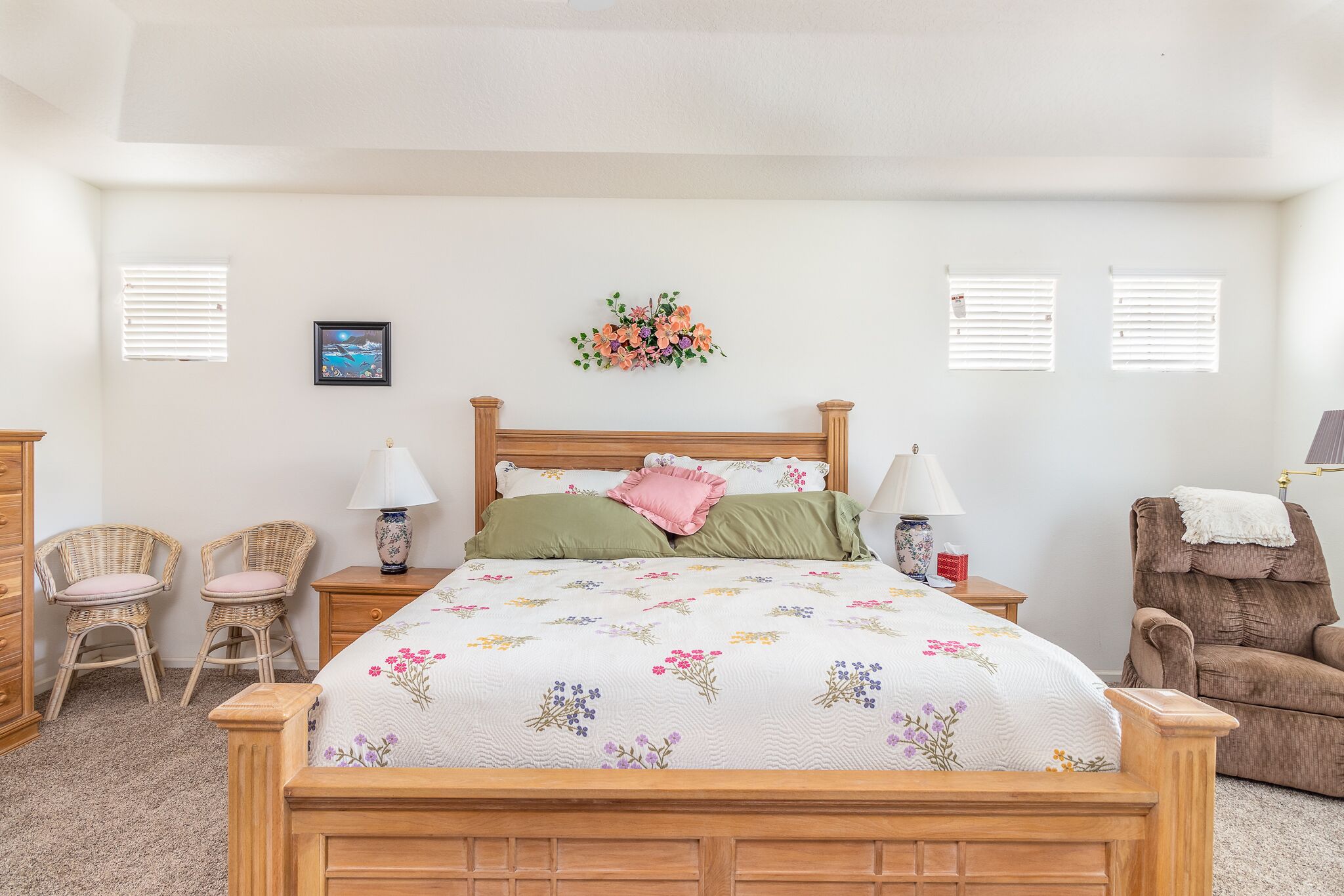 A bedroom with a wooden bed, two wicker chairs, a brown armchair, a nightstand with a lamp, and a wall-mounted floral decoration.
