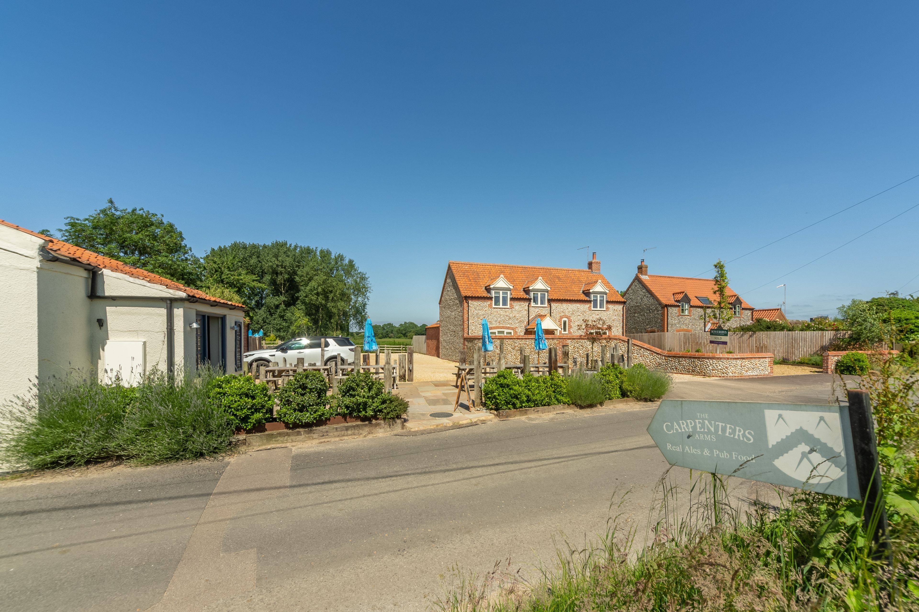 A rural street with a stone building, a white car, and a sign for The Carpenters Arms.
