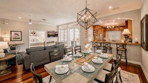 A modern living room with a dining area set for a meal, featuring a chandelier, wooden furniture, and a view of the kitchen.