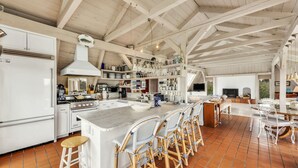 Une cuisine spacieuse avec des meubles blancs, un grand îlot central doté de tabourets, et un coin repas avec un téléviseur et des sièges.