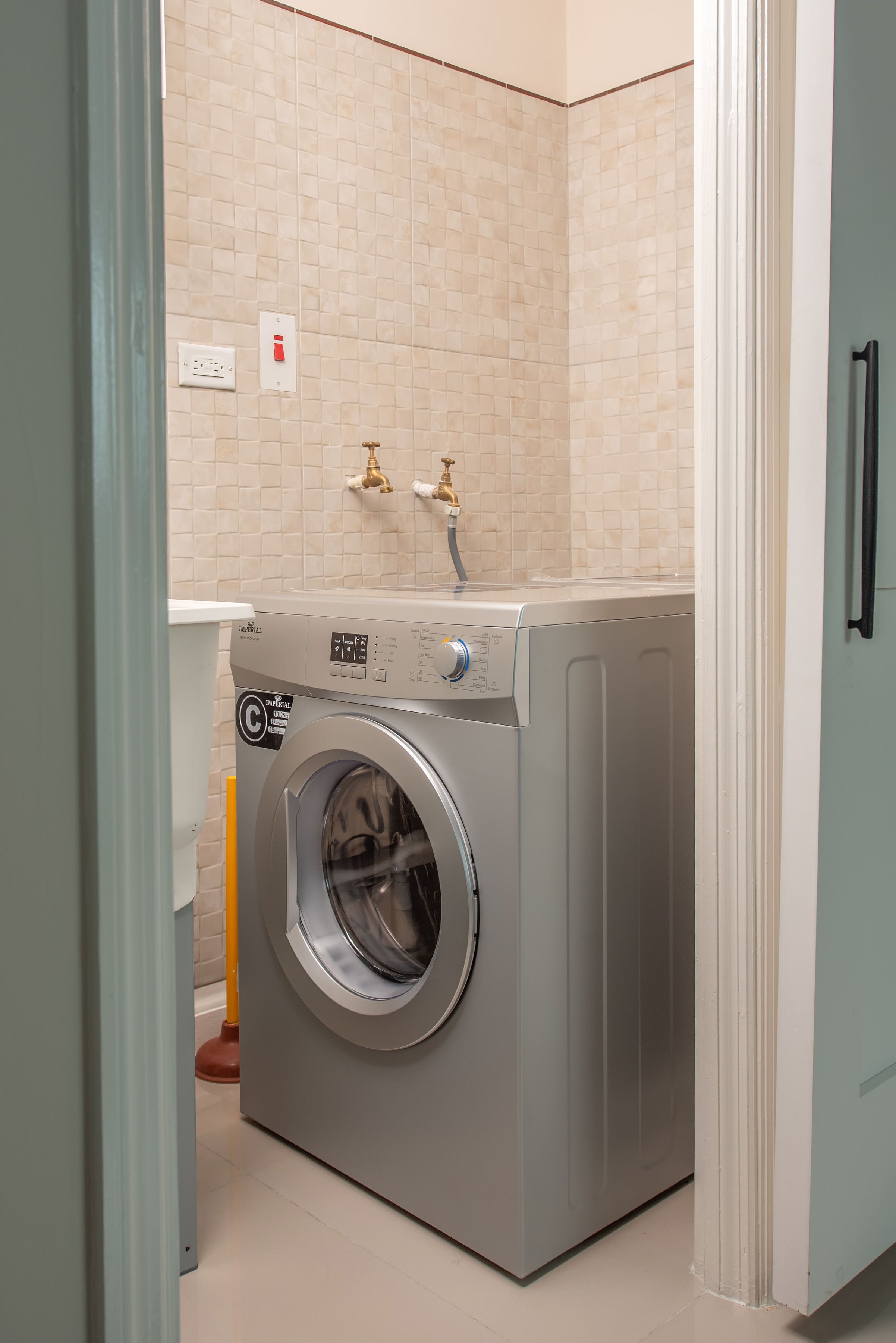 Une petite buanderie équipée d’un lave-linge à chargement frontal, d’un robinet mural et d’un distributeur de papier toilette.