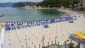 Una spiaggia con file di sedie a sdraio e ombrelloni blu, un pontile di legno e vista sul mare e sulle colline sullo sfondo.