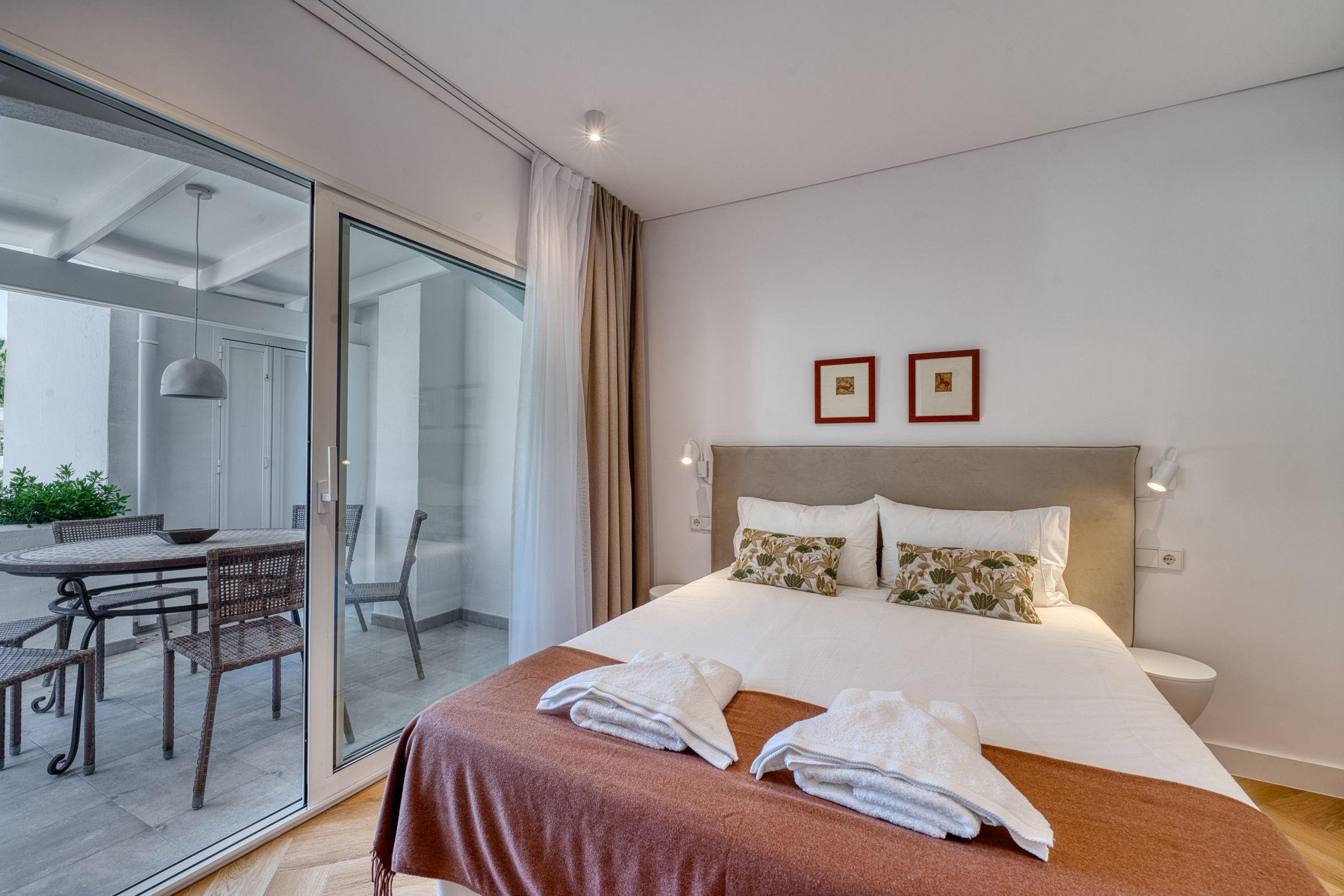 Een moderne hotelkamer met een groot bed, een glazen deur die leidt naar een balkon met buitenmeubilair, en twee ingelijste foto's aan de muur.