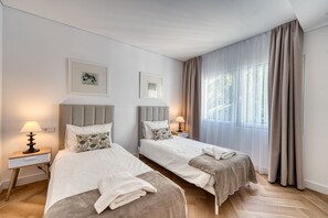 Quarto de hotel com duas camas, uma mesa de cabeceira com luminária e uma janela grande com cortinas.