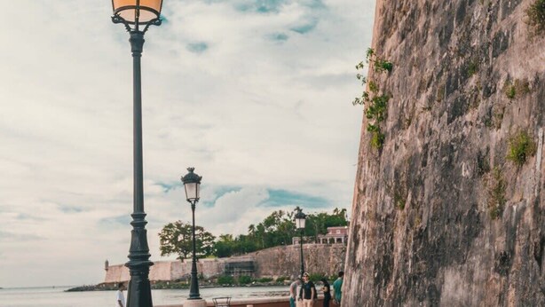 A coastal walkway with stone walls and a tower, featuring a lamp post and a bench.