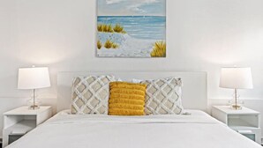 A bedroom with a white bed, a painting of a beach scene, and two bedside lamps.