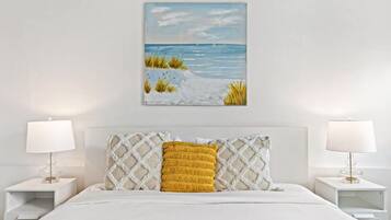 A bedroom with a white bed, a painting of a beach scene, and two bedside lamps.