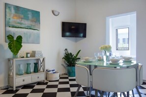 A dining area with a checkered floor, a glass table, white chairs, a flat-screen TV mounted on the wall, and a painting above the table.