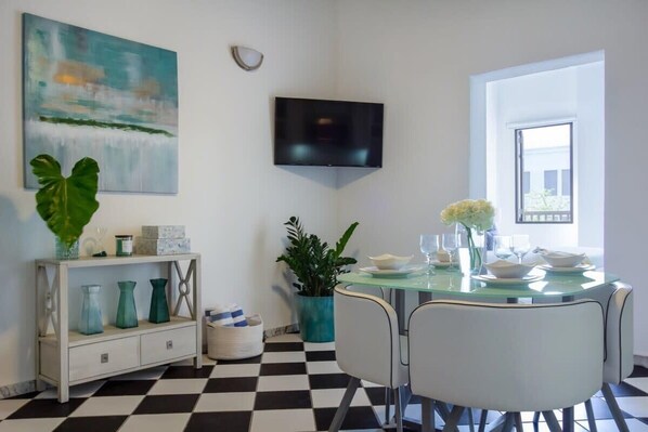 Un comedor con piso a cuadros, una mesa de vidrio, sillas blancas, un televisor de pantalla plana montado en la pared y un cuadro sobre la mesa.