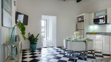 Una cocina compacta con piso a cuadros, gabinetes blancos, microondas y un comedor con mesa y sillas de vidrio.