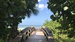 Ein hölzerner Steg führt zu einem Strand, umgeben von üppiger Vegetation und Palmen.