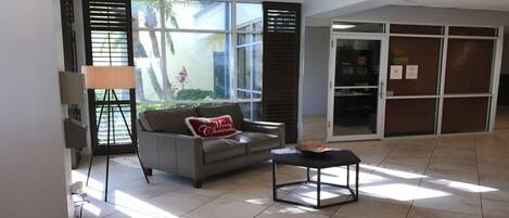 A spacious lobby with a grey sofa, a small table, and a lamp. There are large windows with blinds, and a glass door leading to another room.