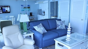 Ein modernes Wohnzimmer mit einem blauen Sofa, weißen Sesseln, einem Glastisch und einem an der Wand montierten Fernseher.