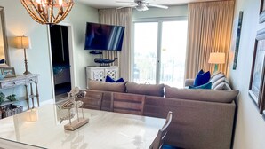 A modern living room with a glass dining table, a sofa, a TV, and a balcony with curtains.