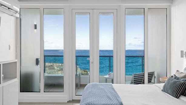 A bedroom with a large bed, a view of the sea through glass doors, and a balcony with a chair and table. - Oceano Studio  | Oceanfront Studio with Kitchen in Trendy Condado Location (San Juan)