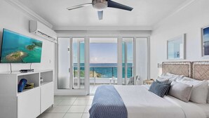A bedroom with a bed, a TV, a ceiling fan, and a view of the ocean through large glass doors. - Oceano Studio  | Oceanfront Studio with Kitchen in Trendy Condado Location (San Juan)